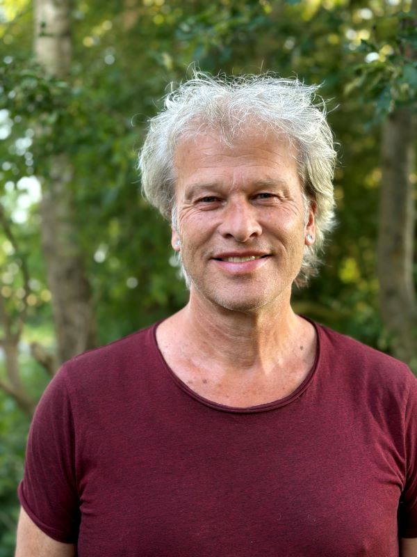 Afbeelding van Leo. Een vijftiger met kort grijzend haar en een vriendelijk gezicht, met groene struiken vaag op de achtergrond.